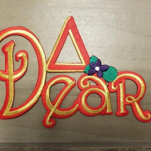 May include: A red and gold embroidered word "Dear" with a small purple and green flower on top.