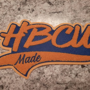 Könnte beinhalten: Ein blauer und orangefarbener Filz-Patch mit dem Text "HBCU Made" in einer stilisierten Schrift.