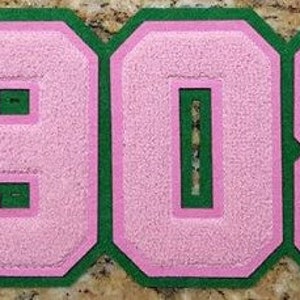 以下が含まれることがあります： 緑色の輪郭を持つピンクのフェルトの数字のパッチで、数字は1908です。