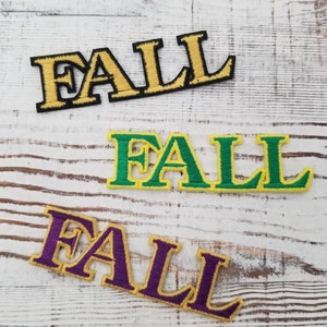 Puede incluir: Tres parches bordados con la palabra "FALL" en una fuente de letras en negrita. Los parches son dorados y negros, verdes y amarillos, y morados y dorados. Los parches están ligeramente inclinados.