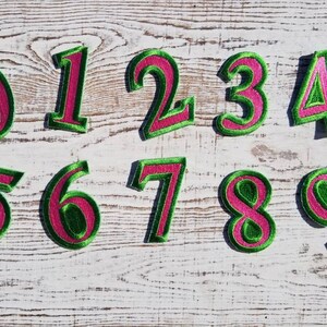 May include: Embroidered number patches, featuring a green border and a pink centre. The numbers 0 to 9 are arranged on a distressed white wooden background. These patches are suitable for various craft projects or customisation.