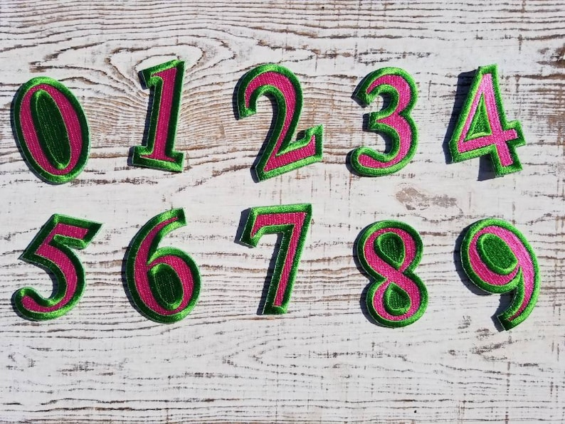 May include: Embroidered number patches, featuring a green border and a pink centre. The numbers 0 to 9 are arranged on a distressed white wooden background. These patches are suitable for various craft projects or customisation.