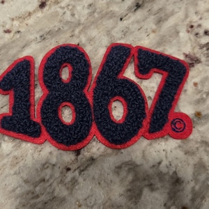 May include: A navy blue and red patch with the number "1867" in a varsity-style font. The numbers are navy blue with a textured appearance and outlined in red. A small copyright symbol is visible.