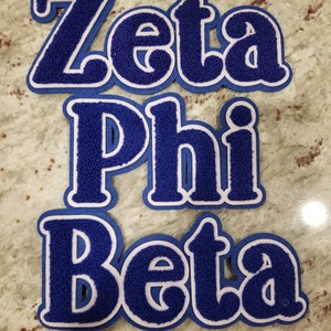May include: Three blue and white Zeta Phi Beta letter patches. The letters are in a bold, collegiate style with a fuzzy texture. Each patch has a white border and a blue backing.