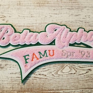 May include: Pink and green felt patch with the text "Beta Alpha" in a cursive font. The patch also has the text "FAMU Spr '93" in a smaller font.