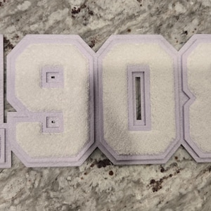 May include: White felt numbers 1908 with a light purple border. The numbers are in a varsity letter style, likely for a jacket or apparel. The background is a speckled gray surface.