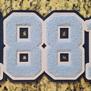 May include: A layered felt patch featuring the number 1881 in light blue chenille with cream borders and a dark navy blue base. The word SPELLMAN is embroidered vertically in dark navy blue thread on the number 1, with a small copyright symbol.
