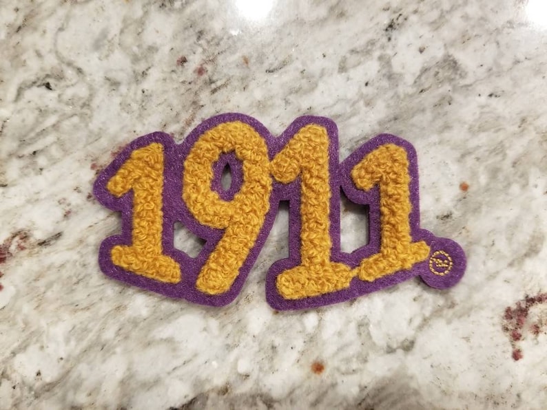 K&ouml;nnte beinhalten: Lilafarbener Filz-Patch mit der Zahl 1911 in gelben, flauschigen Buchstaben.