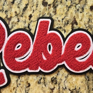 May include: A textured red chenille patch featuring the word Rebels in a cursive script font. The lettering is outlined with a thin white border and a thicker black outer border, creating a layered effect with a soft, fuzzy surface finish.