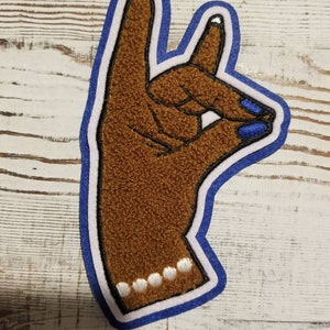 Puede incluir: Un parche de mano de fieltro marrón con un contorno azul y esmalte de uñas azul. La mano está haciendo el signo 'OK' con el pulgar y el índice formando un círculo. La mano tiene una pulsera de cuentas blancas alrededor de la muñeca.