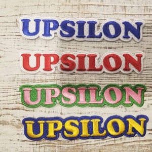 Może przedstawiać: Cztery haftowane łatki z napisem "UPSILON" w różnych kolorach. Łatki są niebieskie, czerwone, różowe i żółte.