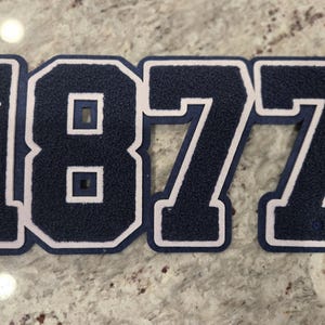 Puede incluir: Un parche de fieltro azul marino con el número "1877" en un estilo de letra universitaria. Los números están delineados en blanco, con el número "1" que también muestra las letras "JSU" verticalmente. El parche es probablemente para una escuela o un equipo.
