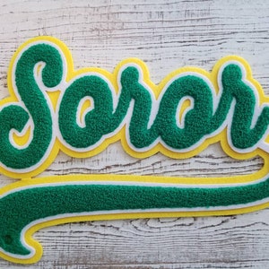 May include: Green and yellow felt applique with the word "Soro" in a cursive font. The applique is shaped like a baseball team logo.