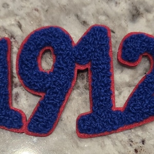 May include: A blue and red patch featuring the number "1912" in a textured, fuzzy fabric. The numbers are blue with a red border. A small red circle with a design is attached to the number "2".