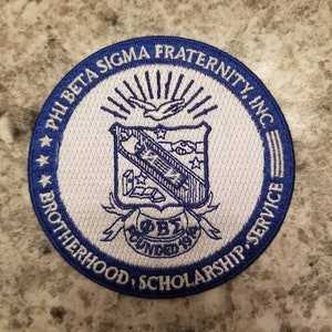 May include: A white and blue embroidered patch with the text "Phi Beta Sigma Fraternity, Inc." and "Brotherhood, Scholarship, Service". The patch also features a crest with a dove and the text "ΦΒΣ Founded 1914".
