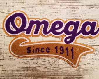 May include: Purple and white felt patch with the word "Omega" in a cursive font. Below the word is a gold felt banner with the text "Since 1911".