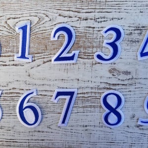 May include: Ten embroidered number patches, each featuring a white centre and a bold blue outline. The numbers 0 to 9 are shown, ideal for personalising clothing or crafts. The patches are arranged on a light-coloured, textured surface.