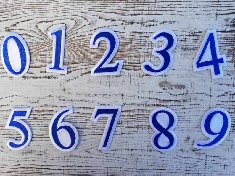 May include: Ten embroidered number patches, each featuring a white centre and a bold blue outline. The numbers 0 to 9 are shown, ideal for personalising clothing or crafts. The patches are arranged on a light-coloured, textured surface.