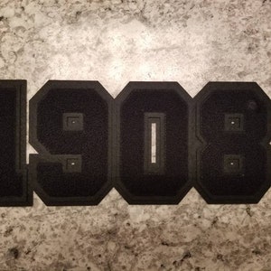 May include: Black felt applique with the number "1908" in a collegiate font. The numbers are outlined in a darker black and have a textured appearance. The applique is likely for a jacket or other garment.