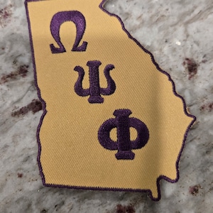 Könnte beinhalten: Ein gold gestickter Aufnäher in Form des Bundesstaates Georgia mit den griechischen Buchstaben Omega, Psi und Phi in Lila.