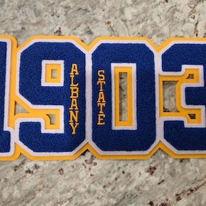 Puede incluir: Un parche de fieltro azul, blanco y amarillo con el número "1903" y las palabras "Albany State" en letras doradas. El parche es probablemente para una escuela o un equipo.