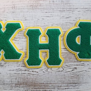 May include: Three felt letters, "X", "H", and "Φ", in green with a yellow outline. The letters are on a white wooden surface.