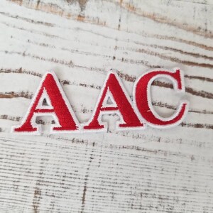 Puede incluir: Tres parches bordados que deletrean "AAC". Cada letra es roja con un borde blanco. Los parches se muestran sobre una superficie de madera pintada de blanco envejecido.