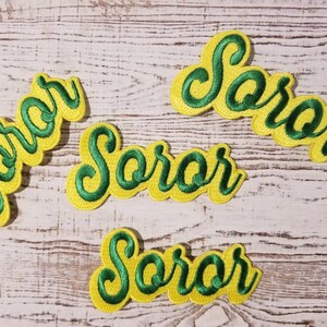 May include: Four yellow and green embroidered patches with the word "Soror" in a cursive font.