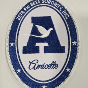 May include: An oval-shaped embroidered patch with a white center and a blue border. The text ZETA PHI BETA SORORITY INC. is arched at the top. A large blue letter A with a white dove silhouette is in the center, with the word Amicette below it.