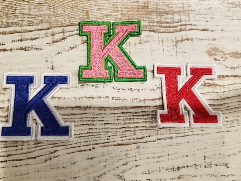 May include: Three embroidered letter "K" patches in different color combinations. One is pink with a green border, another is blue with a white border, and the last is red with a white border. The patches are on a distressed, white-painted wooden surface.
