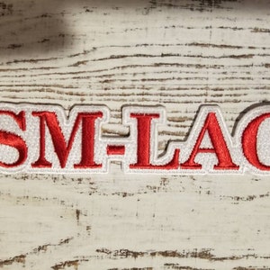 Może przedstawiać: Czerwona naszywka haftowana z tekstem "SM-LAC" w białych obramowanych literach.