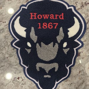 以下が含まれることがあります： 紺色と白のバイソンの形をした刺繍パッチ。パッチには、赤いスクリプトで「Howard」という単語と数字「1867」が書かれています。バイソンは白い角と灰色のディテールを持っています。