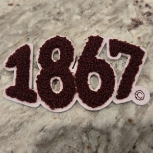 Peut inclure: Écusson bordeaux et blanc avec le chiffre "1867" dans un design texturé et cousu. Les chiffres sont bordeaux avec un bord blanc, sur un fond clair et moucheté. Un petit motif circulaire est visible.