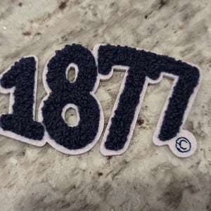 Könnte beinhalten: Ein marineblauer Chenille-Aufnäher mit der Nummer "187" im College-Stil. Die Zahlen sind hellrosa umrandet. Ein kleines Copyright-Symbol ist unten rechts auf dem Aufnäher zu sehen.