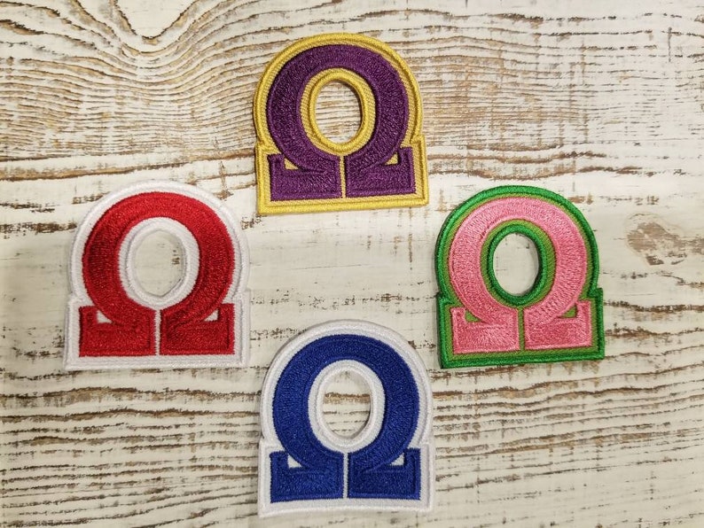 May include: Four embroidered iron-on patches featuring the Greek letter Omega in different colours: red, purple, pink, and blue. Each patch has a white outline and a white base.