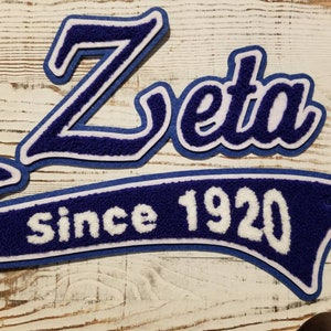 May include: Blue and white Zeta Greek letter patch with the word "Zeta" and the text "Since 1920" on a white wooden background. The patches have a textured, felt-like appearance.