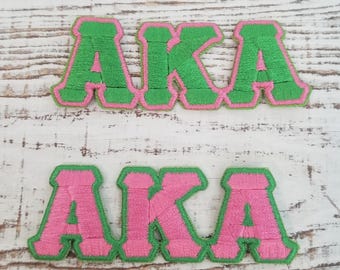 May include: Two embroidered patches with the letters "AKA" in pink and green. The top patch has green letters with a pink outline, and the bottom patch has pink letters with a green outline.