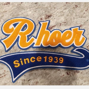 May include: A blue and yellow felt patch with the word "Rhoe" in a cursive font and the text "Since 1939" in a smaller font.