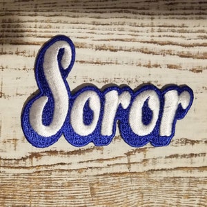 以下が含まれることがあります： 白文字で「Soror」と書かれた刺繍パッチ。青色の縁取りが施されています。パッチは青い縁取りがあり、明るい木目調の背景に配置されています。