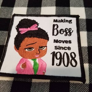 May include: Embroidered patch featuring a cartoon illustration of a Black girl with a pink bow in her hair, wearing a pink jacket and green shirt. The text "Making Boss Moves Since 1908" is embroidered in black on a white background.