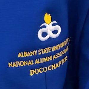 May include: Royal blue jacket with white and yellow emblem. The emblem features a stylized design above the text "ALBANY STATE UNIVERSITY NATIONAL ALUMNI ASSOC DOCO CHAPTER" in gold lettering.