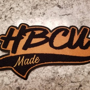 May include: A black and gold patch with the text "HBCU Made" in a varsity-style font. The patch has a black border and a gold-colored center, with the words embroidered in a contrasting color.