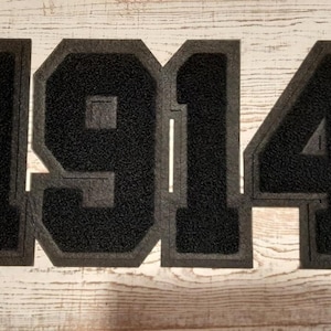 May include: Black felt applique with the number "1914" in a collegiate font. The numbers have a textured appearance and are outlined in a lighter gray. The background is a light-colored, weathered wood.