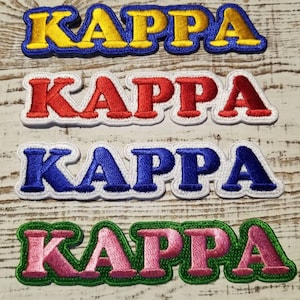 Op de afbeelding: Vier geborduurde strijkpatches met het woord "KAPPA" in verschillende kleuren: geel, rood, blauw en roze. De patches liggen op een houten oppervlak.