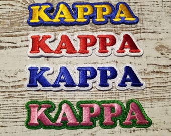 May include: Four embroidered iron-on patches with the word "KAPPA" in different colors: yellow, red, blue, and pink. The patches are on a wooden surface.