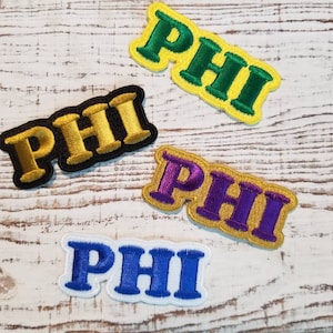 May include: Four embroidered patches with the word "PHI" in different colors: gold, green, purple, and blue. The patches are on a white wooden surface.
