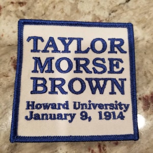 May include: A square, white patch with blue embroidered text and a blue border. The text reads "TAYLOR MORSE BROWN" above "Howard University January 9, 1914".