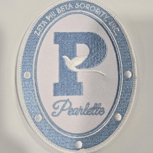 May include: An oval-shaped embroidered patch featuring a light blue border with white circular accents. The center is white with a large light blue letter P, a white dove silhouette, and the text Pearlette. The text Zeta Phi Beta Sorority Inc. is arched along the top.