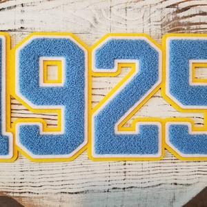 Könnte beinhalten: Ein blauer und gelber Filz-Patch mit der Nummer 1925. Die Zahlen sind weiß und gelb umrandet.