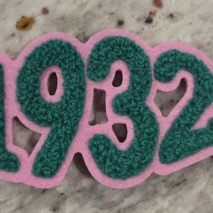 May include: Pink and green felt patch with the number 1932 in a retro style font. The patch has a copyright symbol in the bottom right corner.